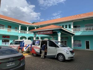A turquoise-colored building with white columns and a red-tiled roof in Roatan, Honduras, features an emergency sign and several parked rescue and medical response vehicles in front.