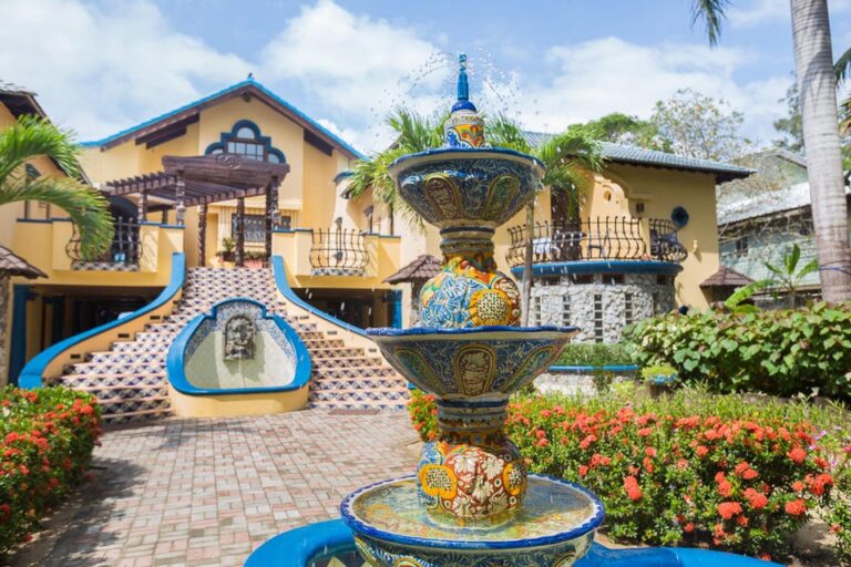 Vibrant ceramic fountain with water spouting at the entrance of a colorful villa in Roatan, Honduras, surrounded by tropical plants and flowers.