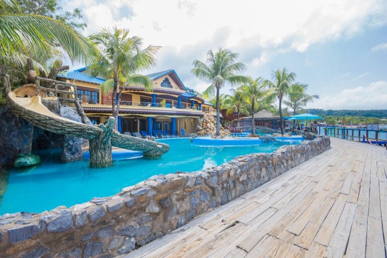Luxurious beachfront resort in Roatan, Honduras featuring a large swimming pool surrounded by palm trees, a wooden deck, and a tropical waterfront view.