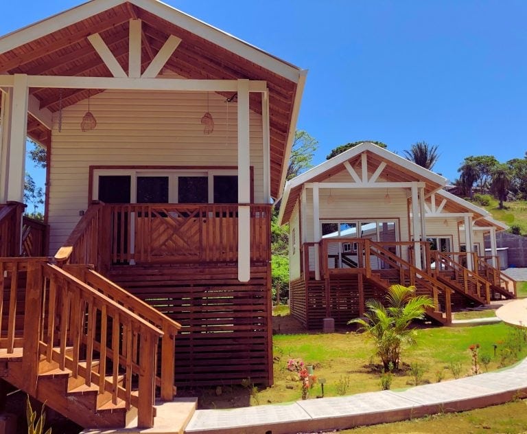 Newly constructed wooden houses with stairs and porches are set against a bright blue sky in Roatan, Honduras, featuring lush greenery and colorful plants.