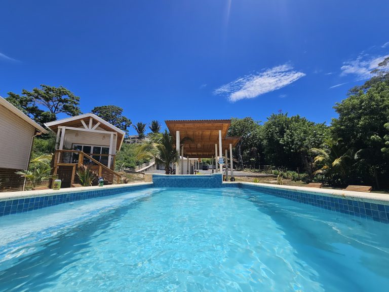 A swimming pool surrounded by white and wooden structures with trees and greenery in the background on Roatan, Honduras.