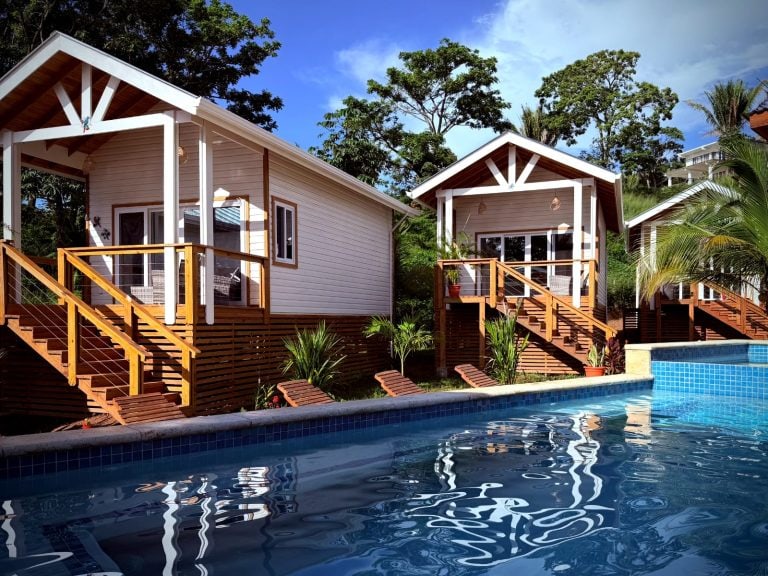 Two white cottages with wooden stairs and railings are situated above a blue swimming pool surrounded by lush green trees on Roatan, Honduras.