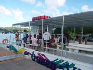 People are boarding a colorful boat with a painted whale design at a dock in Roatan, Honduras, with a covered seating area and scenic hills in the background.