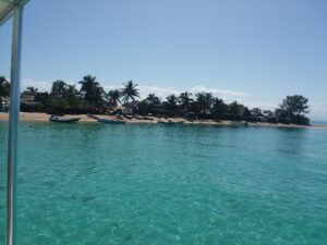 A beachfront scene in Roatan, Honduras, showing turquoise water, several small boats docked along the sandy shore, and a row of palm trees with thatched-roof huts in the background under a clear blue sky.