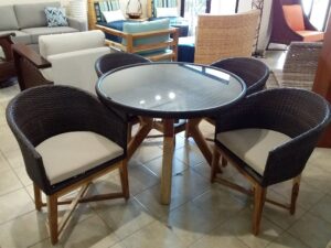 A dining area in Roatan, Honduras features a round glass-top table surrounded by wicker chairs with cream cushions, set on a tiled floor with a variety of colorful and woven furniture and decor in the background.