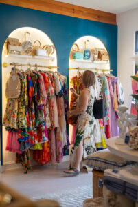 A woman browsing colorful tropical dresses and accessories displayed on racks inside a boutique on Roatan, Honduras, with woven bags and decor on illuminated shelves against a blue wall.
