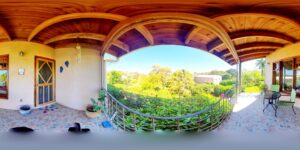 A spacious balcony in Roatan, Honduras, features a wooden arched ceiling, potted plants, a small table with chairs, and views of lush green trees and clear blue sky.