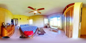 A bright, yellow-walled bedroom in Roatan, Honduras, features a bed with blue and white patterned bedding, a wooden dresser, a ceiling fan, large windows with white curtains, and decorative owl wall hangings. A wooden bench is placed by the window, and a door leads to an adjoining room.