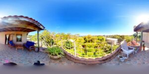 A spacious rooftop terrace in Roatan, Honduras, features outdoor seating with blue and white chairs, potted plants, and a scenic view of lush green trees and the horizon under a clear blue sky.