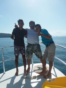Three men are smiling and posing on a boat deck with the ocean and a distant island visible in the background, during daytime in Roatan, Honduras.