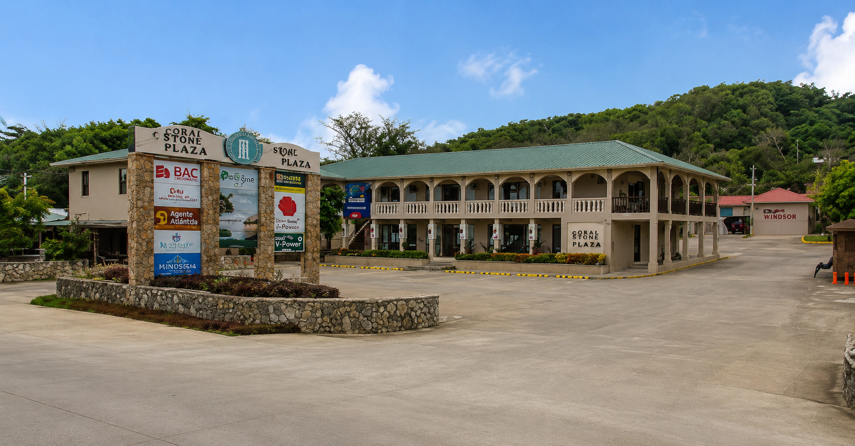 The Coral Stone Plaza in Roatan, Honduras, features a two-story building with a green metal roof and arched verandas, surrounded by lush green hills and a clear blue sky.