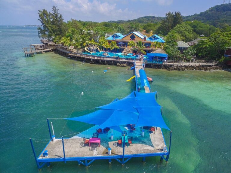 Aerial view of a colorful restaurant and dock extending into clear turquoise waters in Roatan, Honduras, with lush green hills in the background.