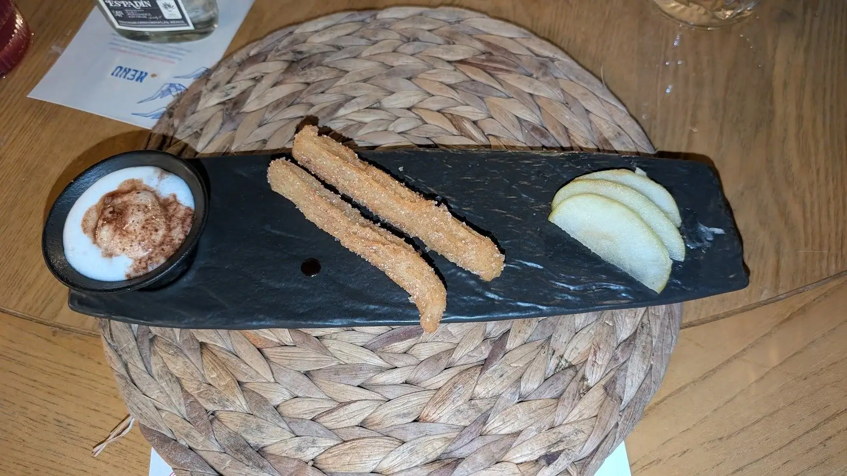 Churros with a side of sliced green apples and a small cup of chocolate sauce on a black slate serving platter, placed on a woven placemat on a wooden table in Roatan, Honduras.