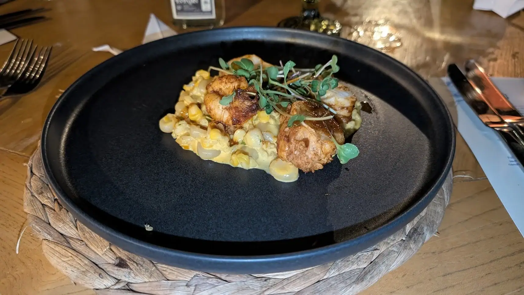 A plated dish of fried chicken served over creamy corn and pasta, garnished with microgreens on a black skillet in a restaurant setting in Roatan, Honduras.