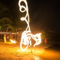 A performer creates intricate fire poi swings on a beach at night in Roatan, Honduras, with palm trees and an audience in the background.