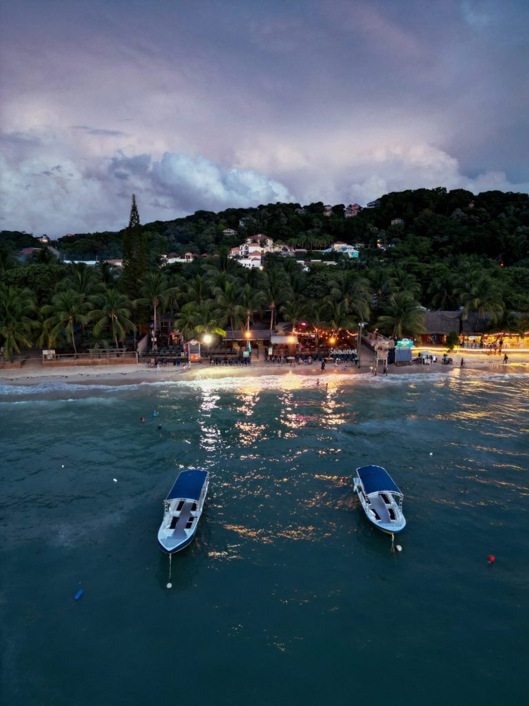 A view of a beachfront bar in Roatan, Honduras, with palm trees, illuminated signs, and buildings on a hillside, surrounded by the Caribbean Sea, with two boats anchored near the shore at dusk.
