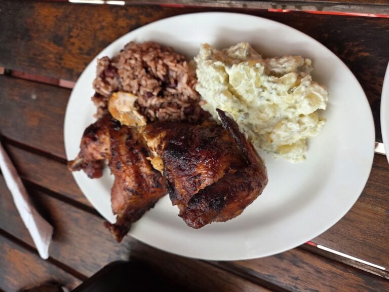 A plate of traditional Honduran cuisine featuring rice and beans, a piece of roasted or grilled meat with sauce, and a serving of potato salad is shown on a wooden table in Roatan, Honduras.
