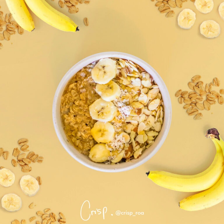 A bowl of oatmeal topped with sliced bananas and chopped nuts is surrounded by scattered oats, dried banana chips, and more bananas on a yellow background in Roatan, Honduras.