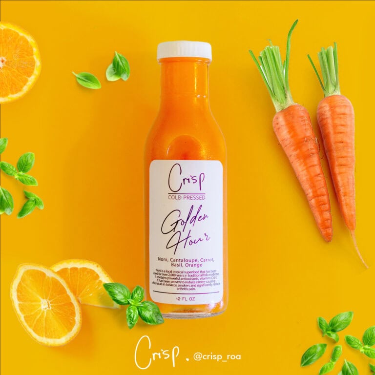A bottle of Crisp Golden Hour juice with ingredients including noni, cantaloupe, carrot, basil, and orange is surrounded by fresh orange slices, carrots with green tops, and basil leaves on a bright yellow background, showcasing a health-focused product from Roatan, Honduras.