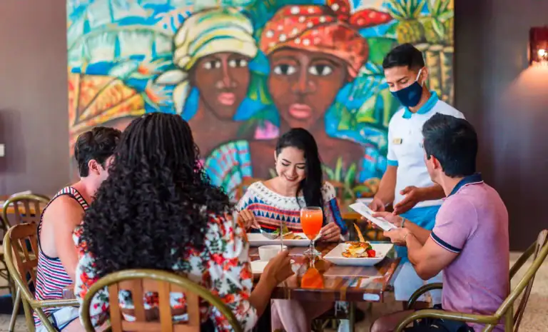 A group of five diverse people dining at a restaurant in Roatan, Bay Islands, Honduras, with a colorful mural of two women wearing traditional headwraps in the background.