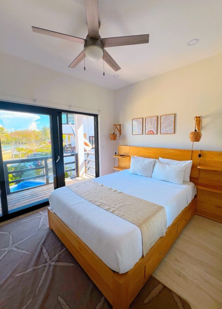 A bedroom in a guesthouse or apartment on Roatan, Honduras, features a wooden bed with white bedding, a bedside shelf, framed wall art, hanging woven lamps, and a large glass sliding door opening onto a balcony with a view of palm trees, a pool, and colorful buildings near the beach.