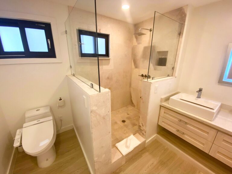A modern bathroom in Roatan, Honduras, features a glass-enclosed shower with a rain showerhead, a white toilet, a small vanity with a sink, and a window with frosted glass for privacy.