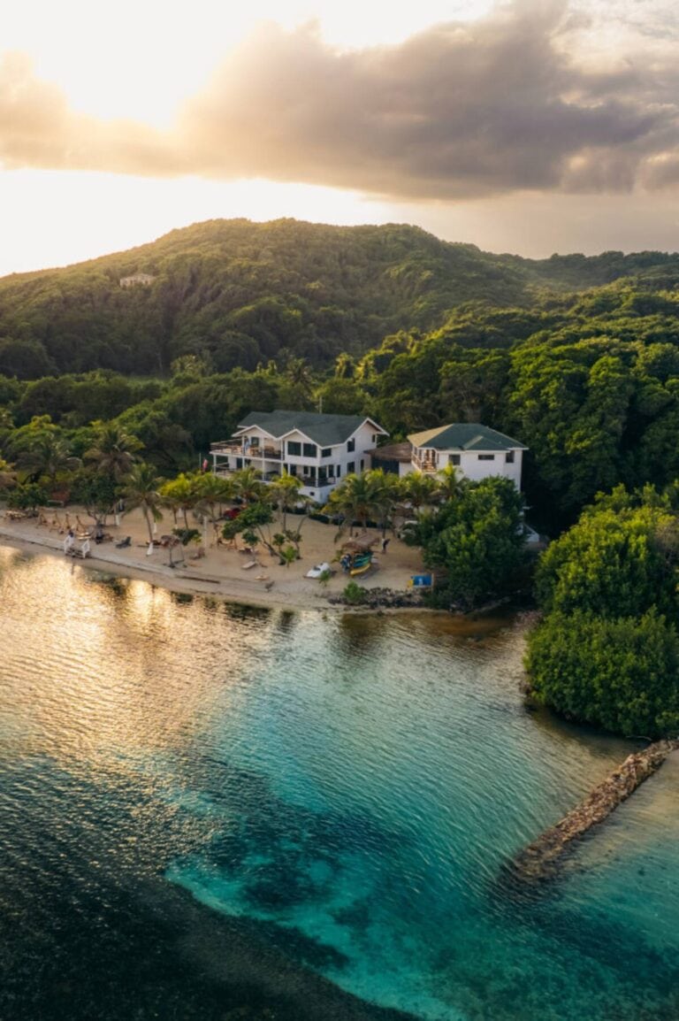 An aerial view of a beachfront property with white buildings, palm trees, and a sandy beach in Roatan, Honduras, surrounded by lush green hills and clear blue waters.