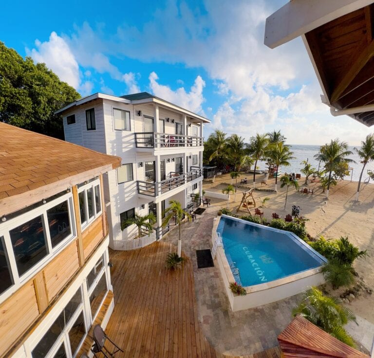 A view from an elevated perspective shows a multi-story white building with a wooden balcony overlooking a sandy beach with palm trees and lounge chairs, a swimming pool, and the ocean shoreline in Roatan, Honduras under a blue sky with scattered clouds.