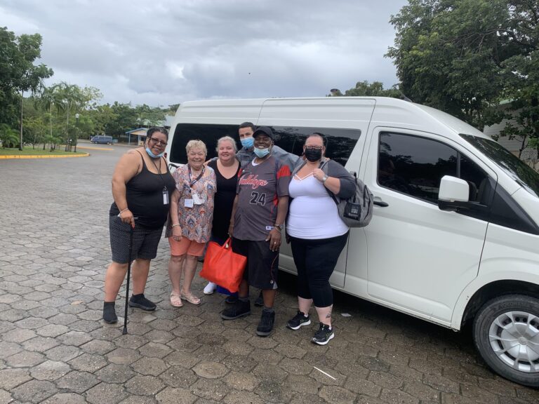 A group of six diverse people wearing masks stands next to a white van in a paved parking area surrounded by lush green trees on Roatan, Honduras, under a cloudy sky.