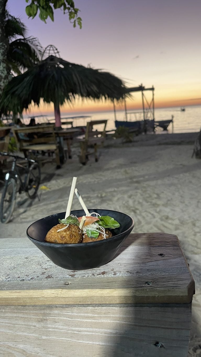 A bowl of fried food garnished with herbs and vegetables is placed on a wooden table at a beachside setting in Roatan, Honduras, with a thatched umbrella, boats on the calm sea, and a sunset sky in the background.