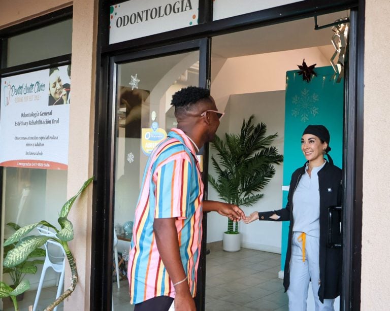 A man in a colorful striped shirt shaking hands with a smiling female staff member wearing a uniform and black cap inside a dental clinic labeled "Odontologia" on Roatan, Honduras.