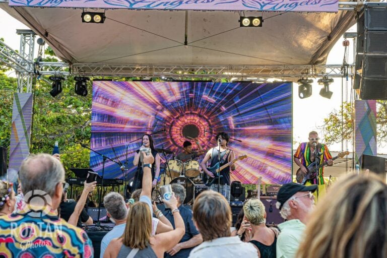 A live band performs on an outdoor stage in Roatan, Honduras, with a large colorful LED screen displaying a vibrant, swirling pattern behind them, as an audience watches and takes photos during daylight.