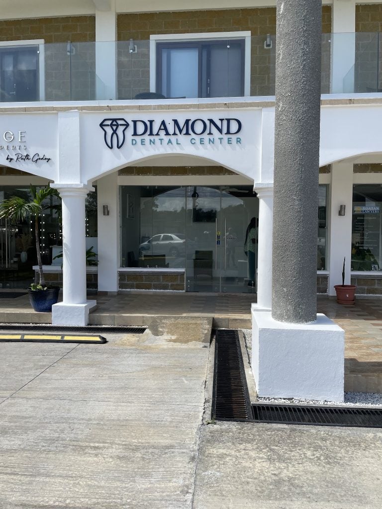 The image shows the storefront of the Diamond Dental Center on Roatan, Bay Islands, Honduras, featuring a white facade with blue signage, glass doors, potted plants, and a parking lot with a drainage grate in front.