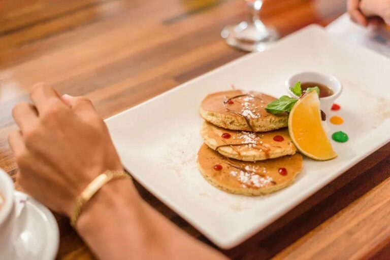 Three golden pancakes topped with powdered sugar, served with a lemon wedge, colorful syrup drops, and a small bowl of syrup on a white rectangular plate in a restaurant setting in Roatan, Honduras.