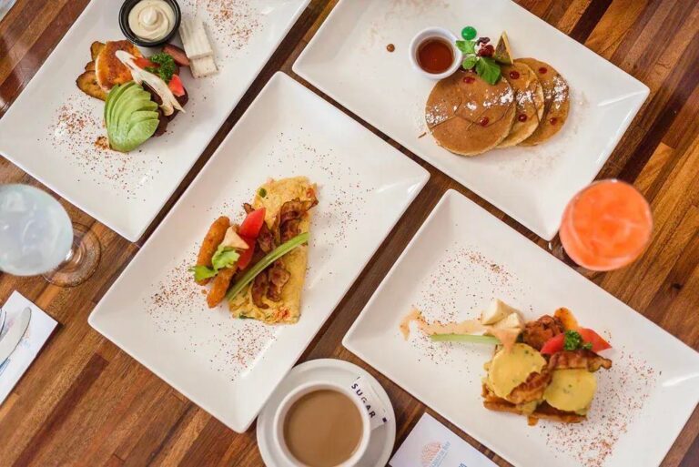 A variety of American and international breakfast dishes served on white plates at a restaurant in Roatan, Honduras, including pancakes with syrup, an omelet with vegetables and bacon, avocado toast with smoked salmon, and a breakfast sandwich with eggs and bacon, accompanied by coffee and fruit drinks on a wooden table.