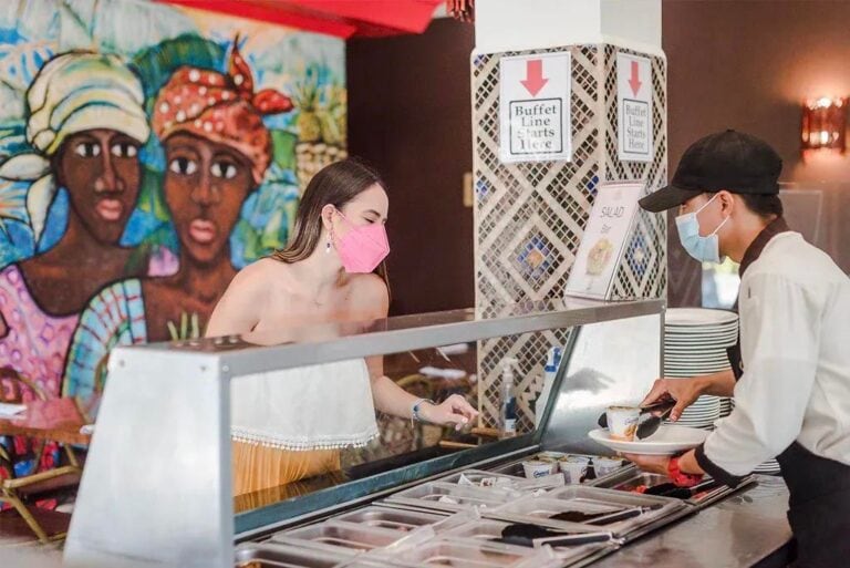 A woman in a pink face mask orders food at a buffet line in a restaurant on Roatan, Honduras, with colorful artwork of local women in traditional attire in the background.