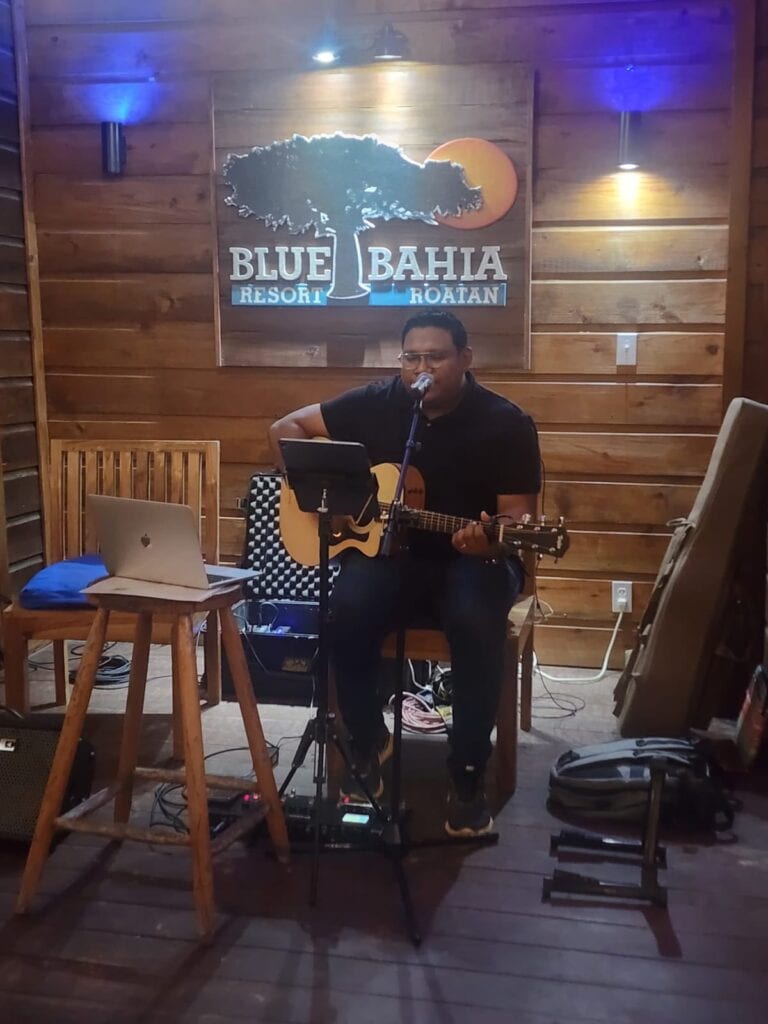 A man performs live music with a guitar and singing into a microphone at Blue Bahia Resort in Roatan, Honduras, with a wooden-paneled wall, a sign displaying the resort's logo, and ambient lighting in the background.