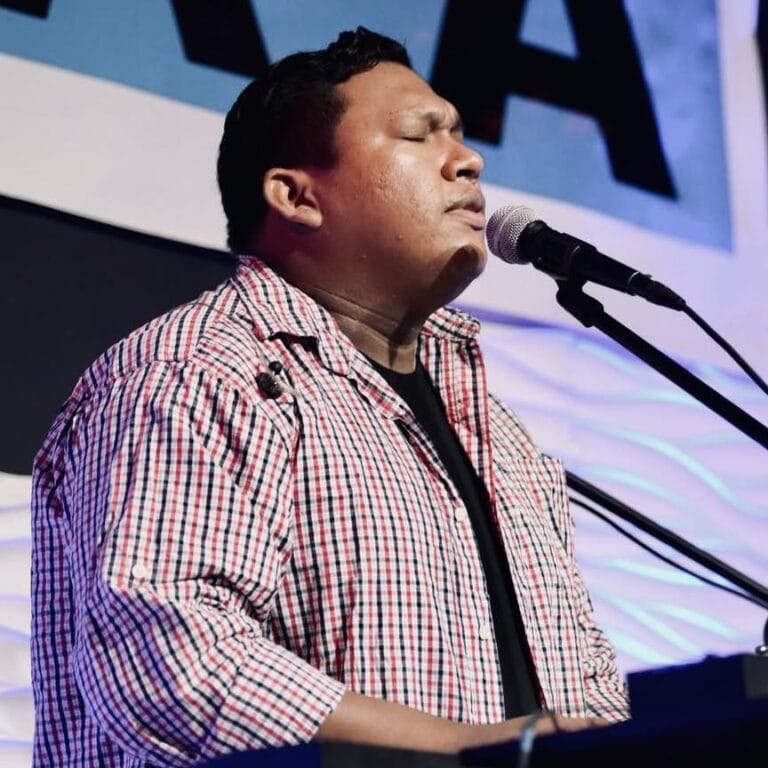 A man with closed eyes and a focused expression performs at a microphone on a stage with a colorful background in Roatan, Honduras.