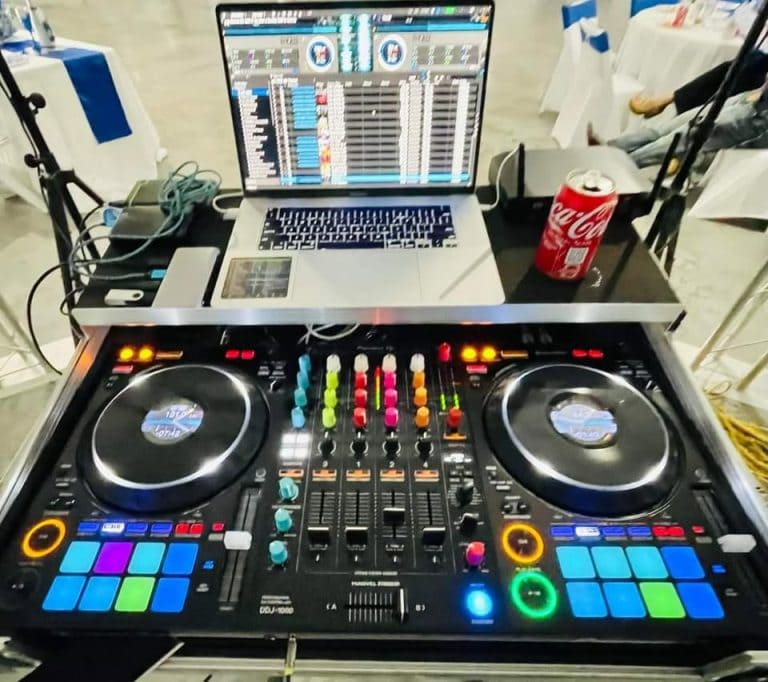 A DJ setup featuring a laptop running music software, a Coca-Cola can, and professional DJ controllers with colorful buttons and knobs, situated on a table at an event in Roatan, Honduras.