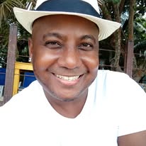 A smiling man wearing a white hat is pictured outdoors on Roatan, Bay Islands, Honduras, with colorful playground equipment and lush greenery in the background.