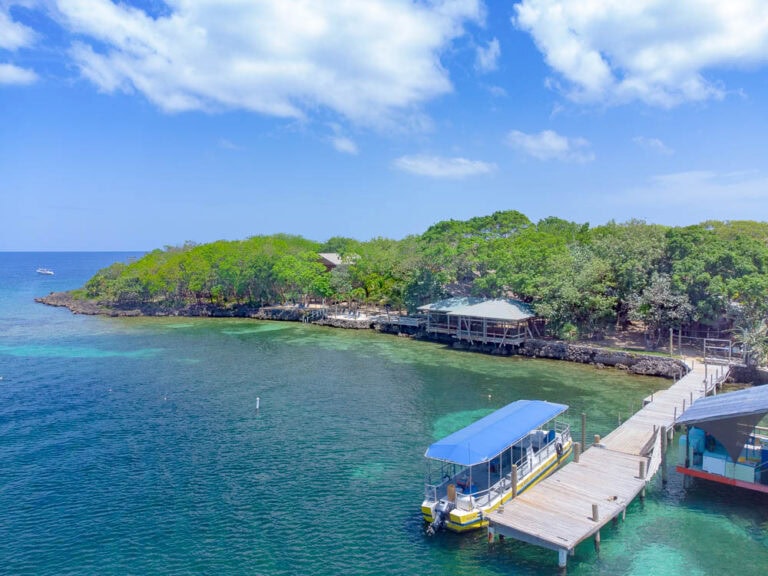 A small harbor in Roatan, Honduras featuring a pier with boats and a lush green island with trees and a wooden structure, under a partly cloudy blue sky.