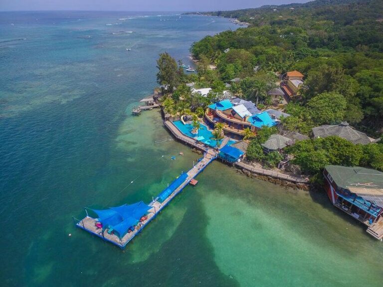 Aerial view of a tropical resort on Roatan, Honduras, featuring a large wooden pier extending into clear blue Caribbean waters with a shaded pavilion at the end, surrounded by lush greenery and beachfront accommodations on the island of Roatan.