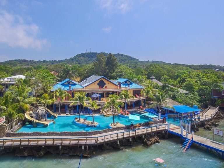 Colorful tropical water park with slides and swimming pools overlooking Roatan, Bay Islands, Honduras, set against lush green hills.
