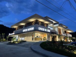 A modern two-story building with illuminated signage reading "Espresso Americano" and "Charli" on Roatan, Honduras, is shown during dusk with a darkening sky and a landscaped parking lot in front.