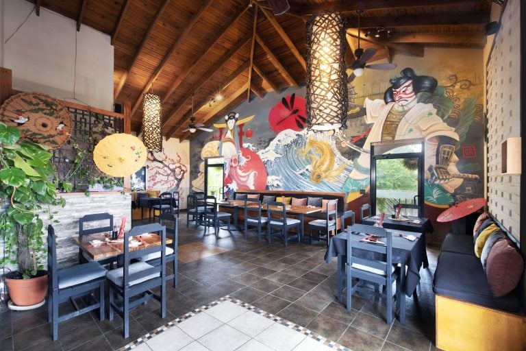 A dining area in a restaurant on Roatan, Honduras, features black tables and chairs, a colorful Japanese-inspired mural with wave, koi fish, geisha, and cherry blossom designs, wooden ceiling beams, decorative paper umbrellas, and Asian-style lanterns.