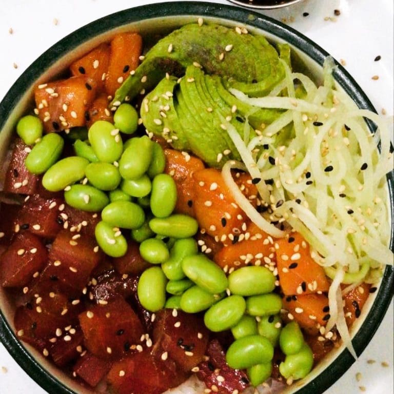 A colorful poke bowl featuring diced raw tuna, edamame, sliced avocado, shredded cabbage, and sesame seeds sitting on a white surface in Roatan, Honduras.