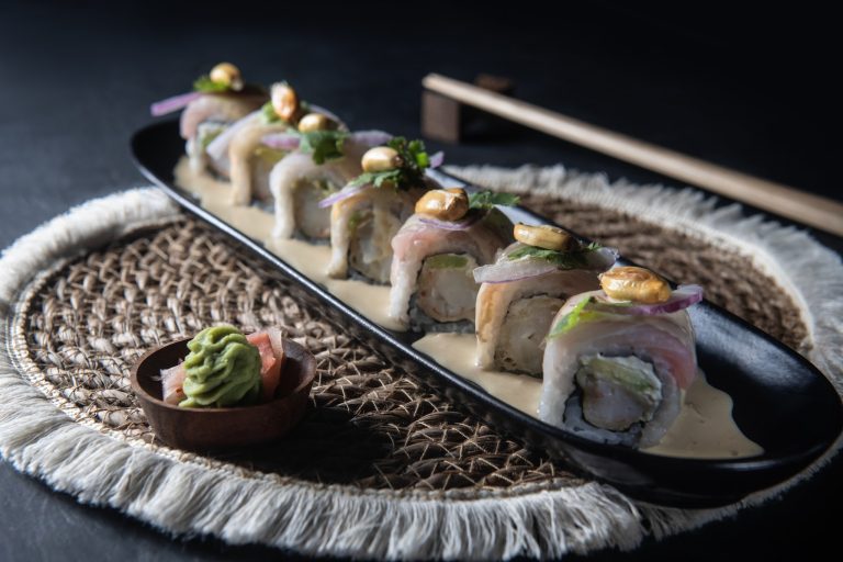 A sushi roll with fresh fish, avocado, and toppings such as cashews and herbs served on a black rectangular plate with a small dish of wasabi and pickled ginger on a woven mat, set against a dark background in Roatan, Honduras.