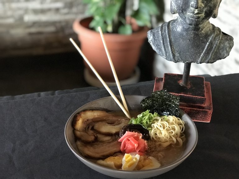 A bowl of ramen with slices of pork, boiled egg, pickled ginger, green onions, and noodles is presented alongside a small sculpture and a potted plant on a dark surface in Roatan, Honduras.