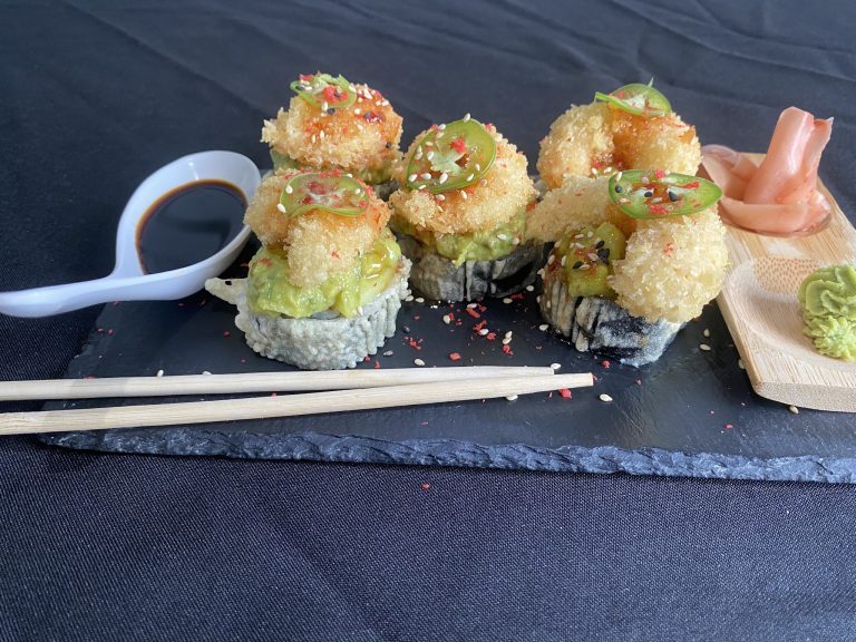 Sushi rolls topped with avocado, spicy sauce, sesame seeds, and chili flakes are presented on a black slate platter with a small dish of soy sauce and a side of pickled ginger in a business setting in Roatan, Honduras.