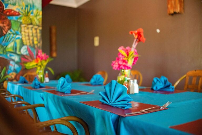 A vibrant restaurant table setup on Roatan in the Bay Islands of Honduras, featuring a turquoise tablecloth, folded blue napkins, and colorful tropical flower centerpieces with lush greenery in the background.